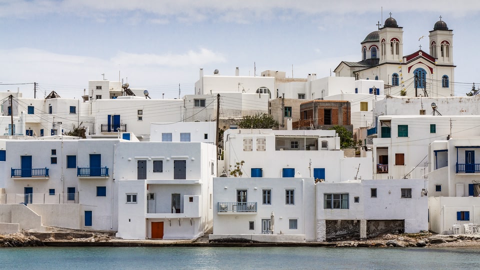 A panoramic view of a cluster of white buildings with blue shutters set against a clear blue sky and overlooking a body of water.