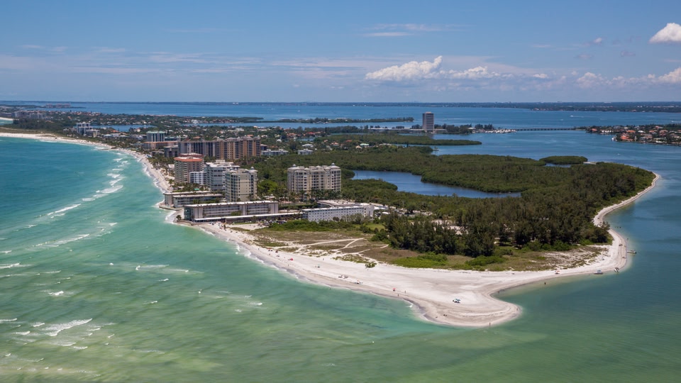 Aerial view of a beach with white sand, turquoise waters, and modern buildings along the shoreline in Sarasota, Florida.