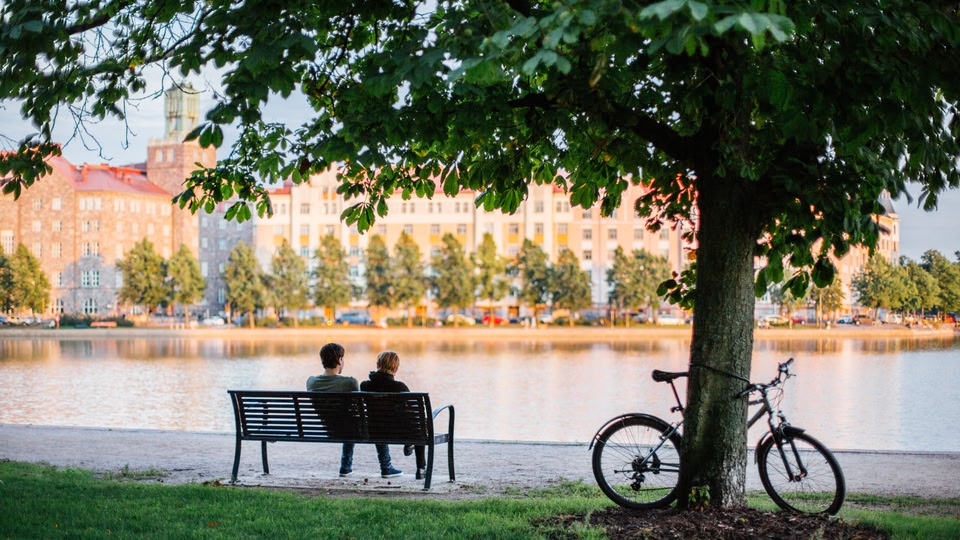 Two people sitting on a bench in a park by the lake with a bicycle leaning against a tree.