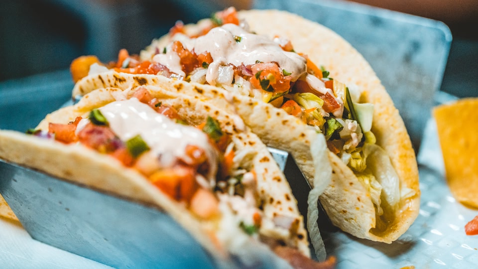Three tacos with creamy white sauce and fresh vegetables served on a metal tray.