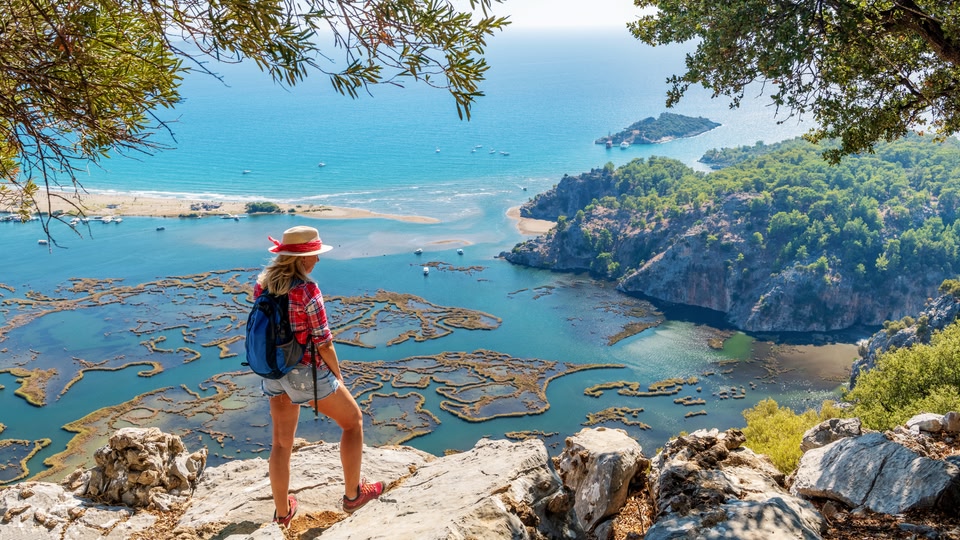 A woman with a backpack stands on a rocky outcrop overlooking a coastal landscape with a lagoon and open sea in the background.