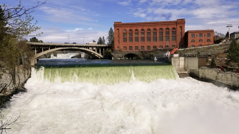 A large waterfall with water cascading over a dam, surrounded by trees and an old brick building in the background.