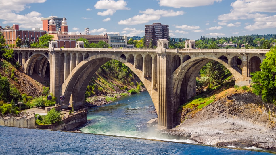 A large stone bridge with multiple arches spans over a flowing river in an urban area, with greenery and buildings visible in the background.