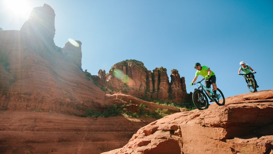 A woman stands atop a red rock formation, overlooking a vast, lush valley with red rock formations and green vegetation in Sedona, Arizona.