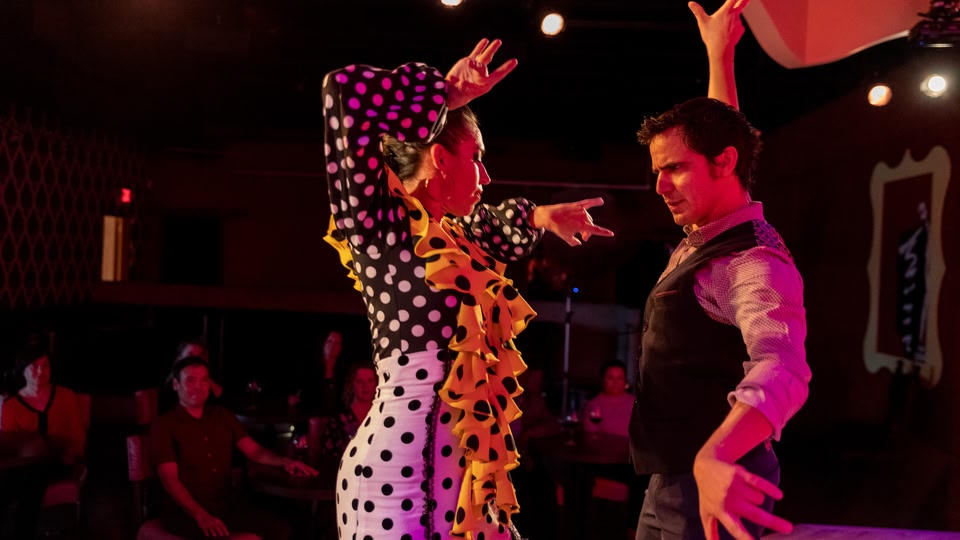 A couple dancing passionately on a stage in a dimly lit indoor venue with other patrons watching from tables.