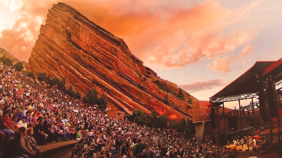 A large crowd of people is gathered outdoors in front of a red rock formation, watching a live concert at sunset.