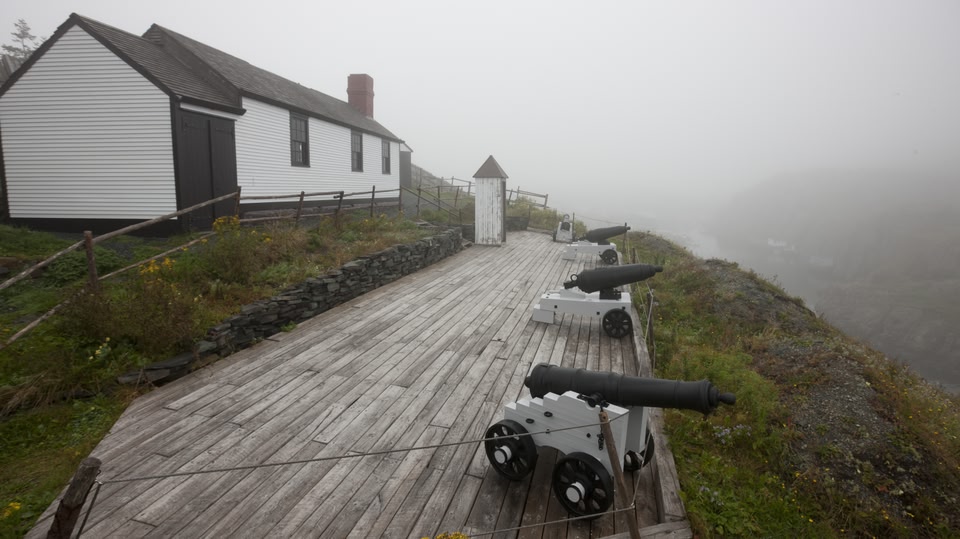 An old white house and several vintage cannons on a wooden platform near a foggy coastline.
