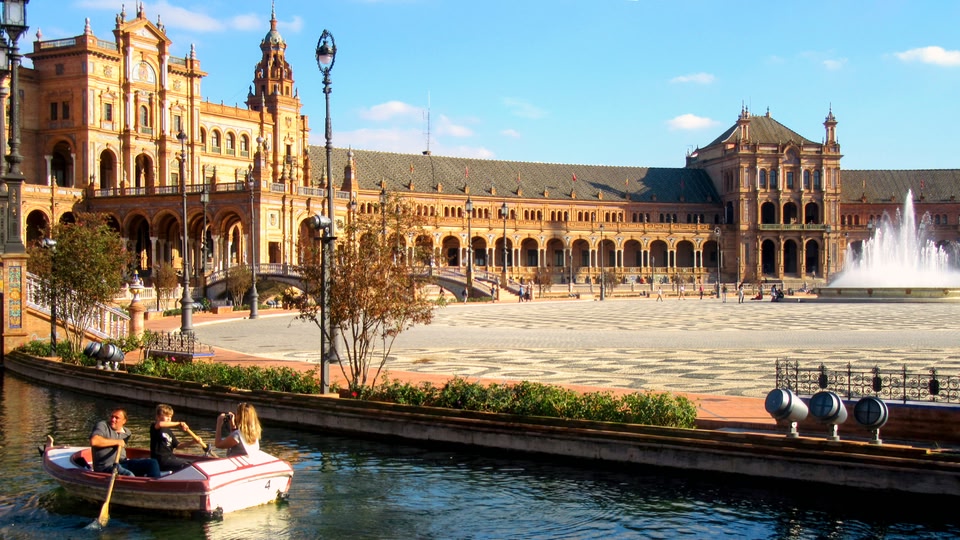A view of the Plaza de España in Seville, Spain, with a boat carrying tourists on a canal in the foreground and the grand architectural structure with a central fountain in the background.