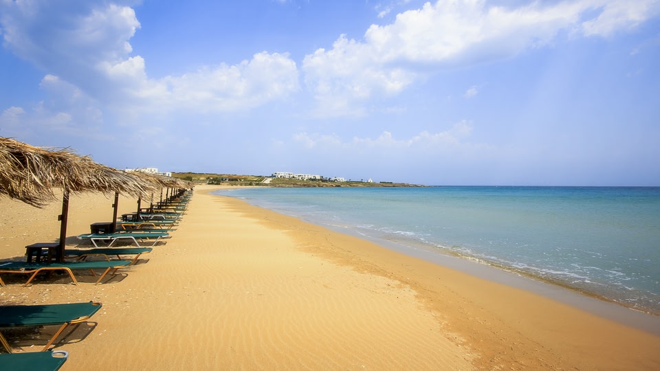 A serene beach scene with green lounge chairs lined up on the sand, facing a calm blue sea under a partly cloudy sky.