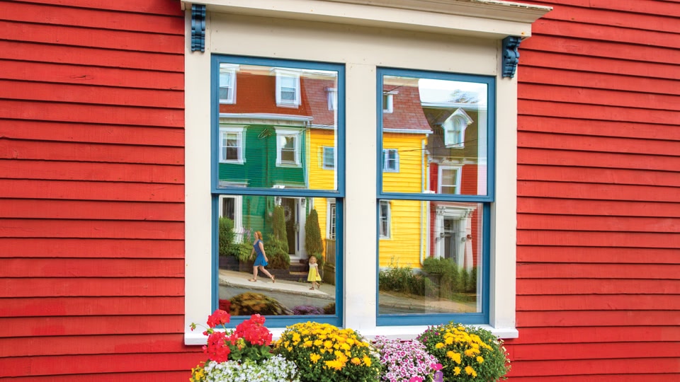 A red house in St John's, Newfoundland and Labrador, Canada, with a window showing a colorful reflection of neighboring houses and people walking on the sidewalk.