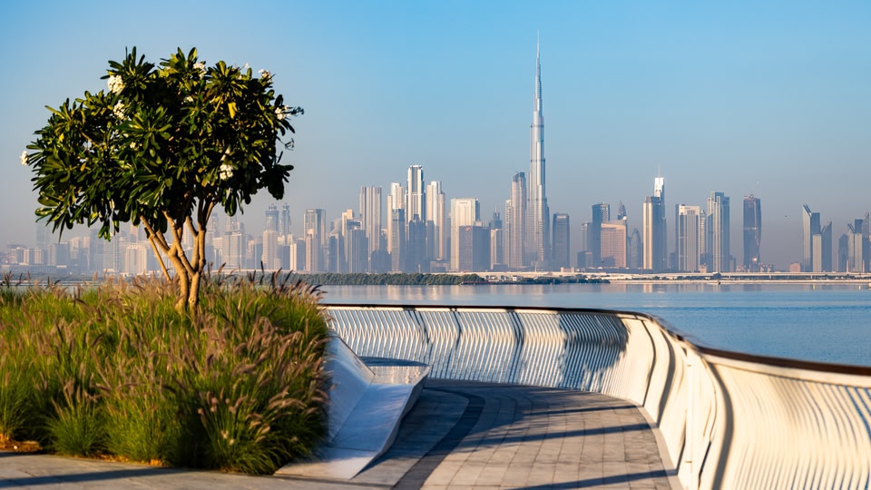 A scenic view of the Dubai skyline with the Burj Khalifa towering in the distance, taken from a waterfront promenade.