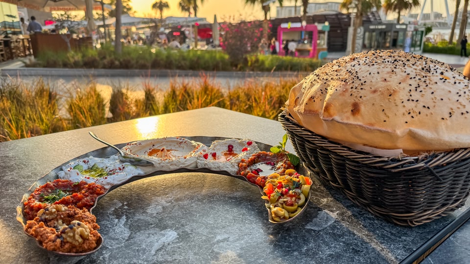 A plate of various Middle Eastern dishes with a basket of bread on a table outdoors.