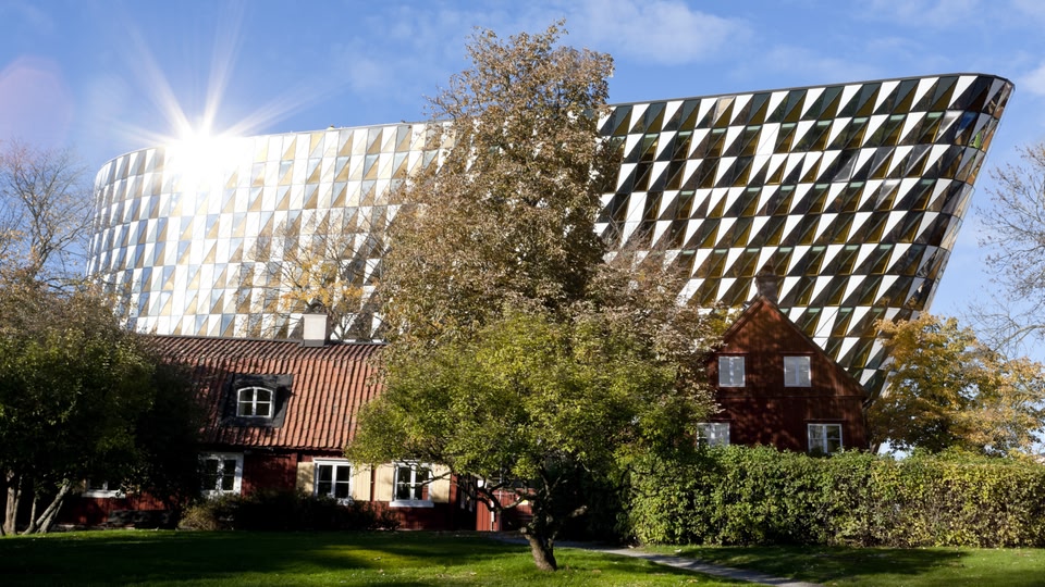 A modern, geometrically designed building with a reflective facade stands next to a traditional red house with white windows. Trees and a hedge are in the foreground.