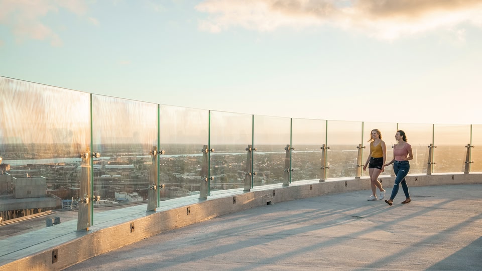 Two women walking on a rooftop with a skyline view during sunset.