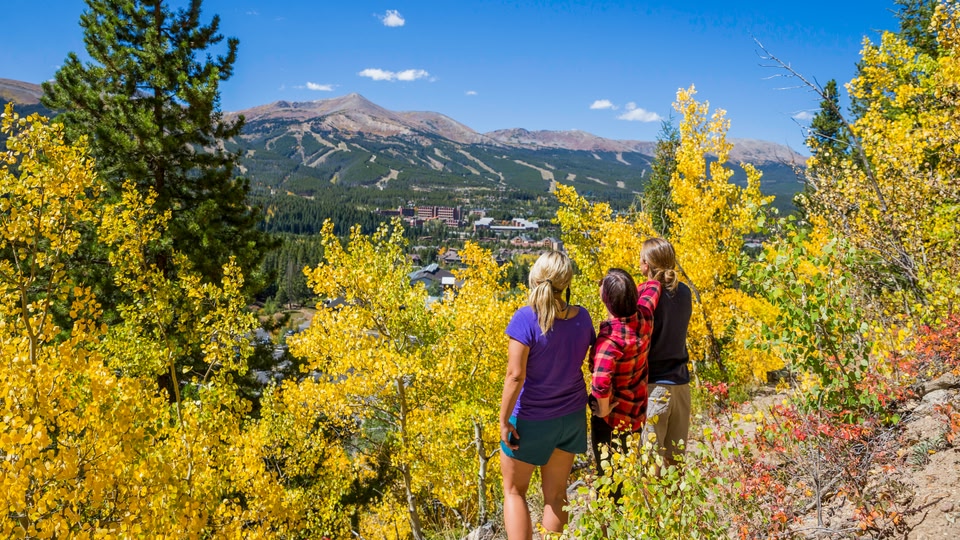 Three people standing on a hillside overlooking a valley with autumn-colored trees and distant mountains.