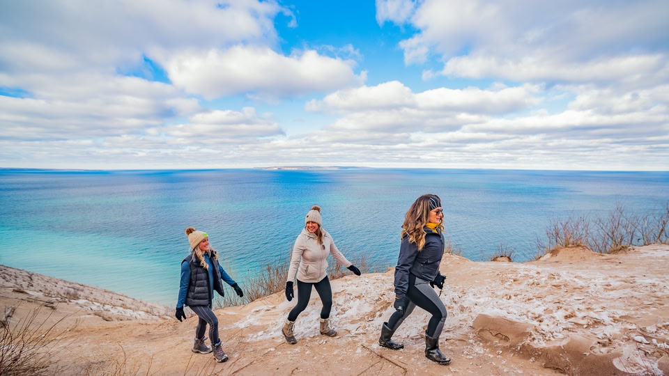 Three people walking along a snowy path near a beach, with a vast blue sea in the background.