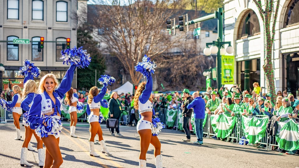 Cheerleaders perform a routine on a city street during a festival, with a crowd of spectators in the background.