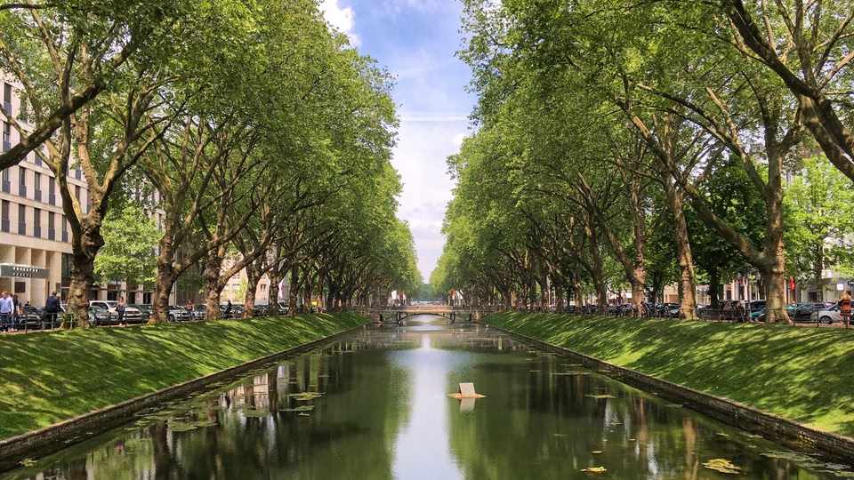 A serene canal in an urban park lined with trees and greenery, with people walking along the sides.
