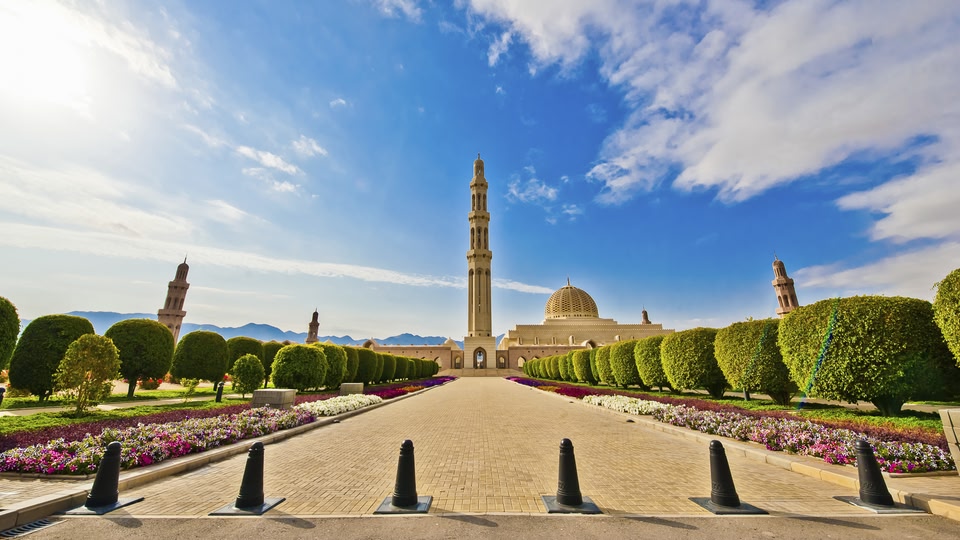 A scenic pathway lined with hedges and flowers leading to a large mosque with a prominent dome and minarets in Muscat, Oman.