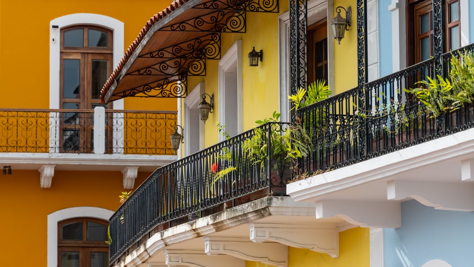Ornate wrought-iron balcony with potted ferns and yellow facades on a colonial building in Panama City.