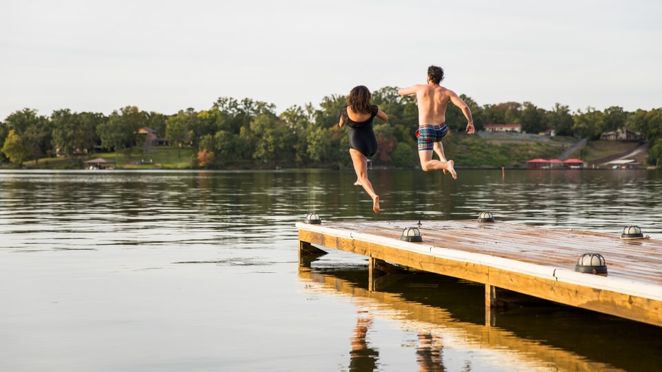 A man and a woman jumping off a wooden dock into a lake during the summer