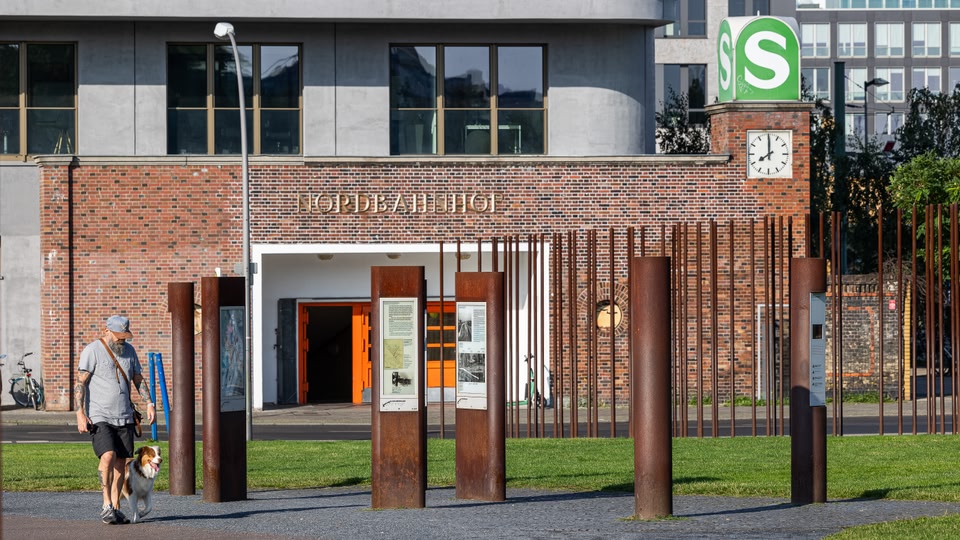 A man with a beard walks his dog past a brick building with an orange door and a clock tower, with vertical iron bars in front of the entrance.