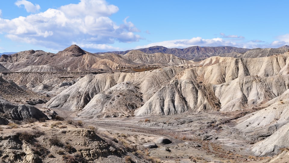 A vast, arid landscape with rugged mountains and rocky terrain under a partly cloudy sky.