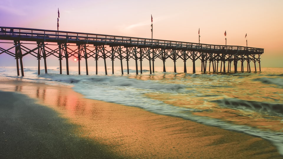 A scenic beach view with a pier stretching out over the ocean, waves crashing onto the shore, and the sun setting in the background with warm orange and pink hues.