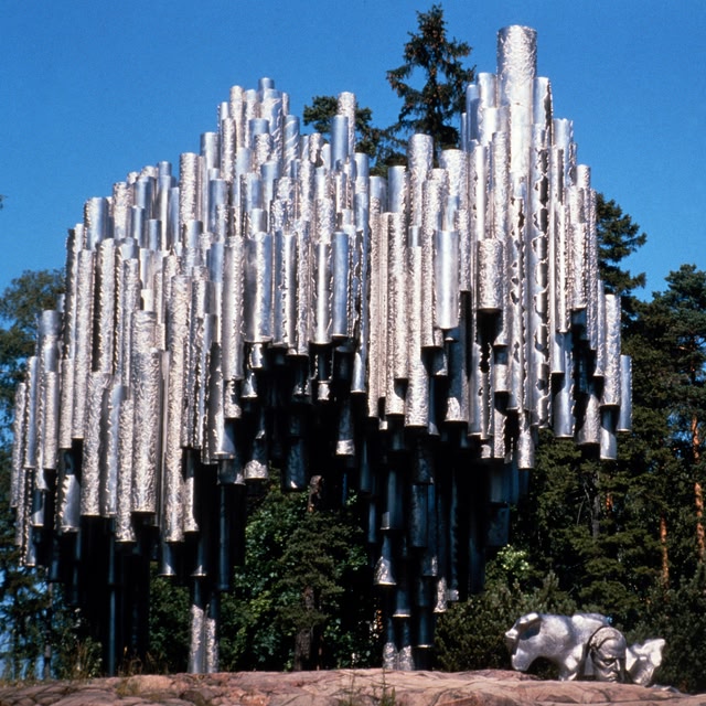 A large, modern sculpture made of silver-colored tubes arranged vertically, situated outdoors with a blue sky and trees in the background.