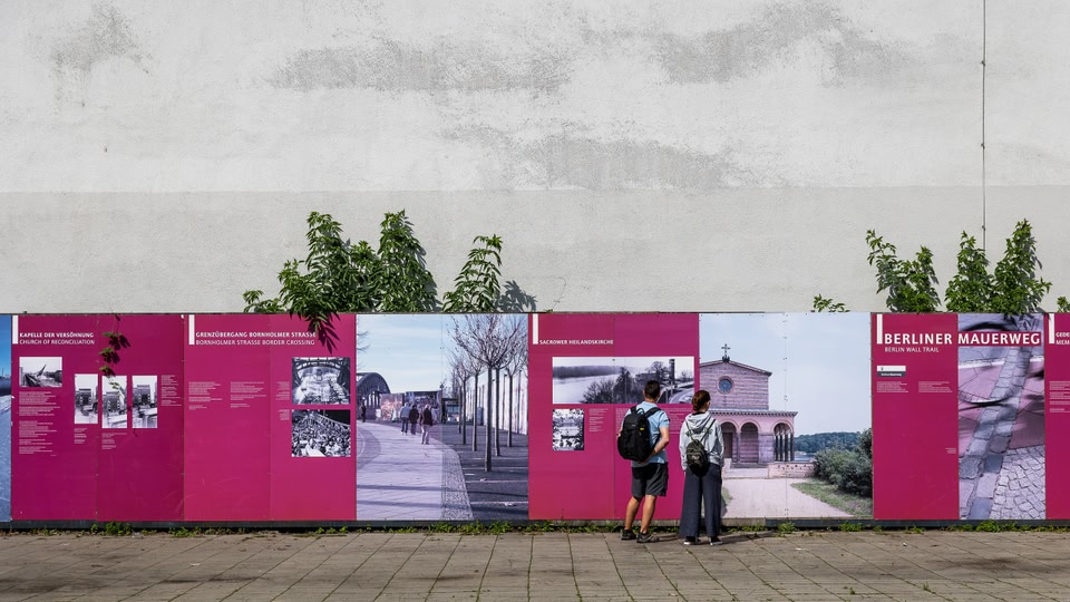 Two people standing in front of a mural depicting historical scenes and buildings at Checkpoint Charlie in Berlin.