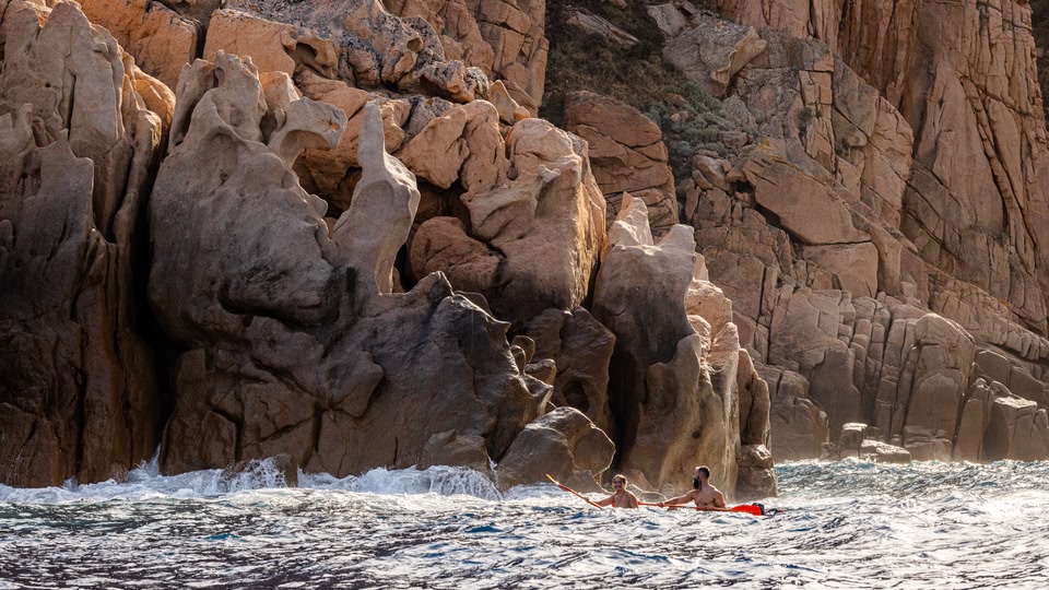 Two people paddling a kayak near weathered granite cliffs at Calanques de Piana, waves breaking at the rock base.