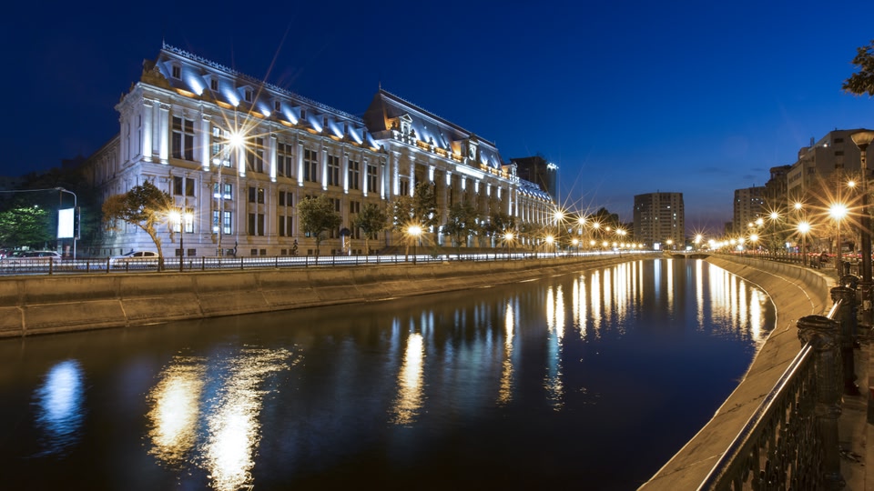 Illuminated city skyline at night with a prominent historic building and a water canal in the foreground.