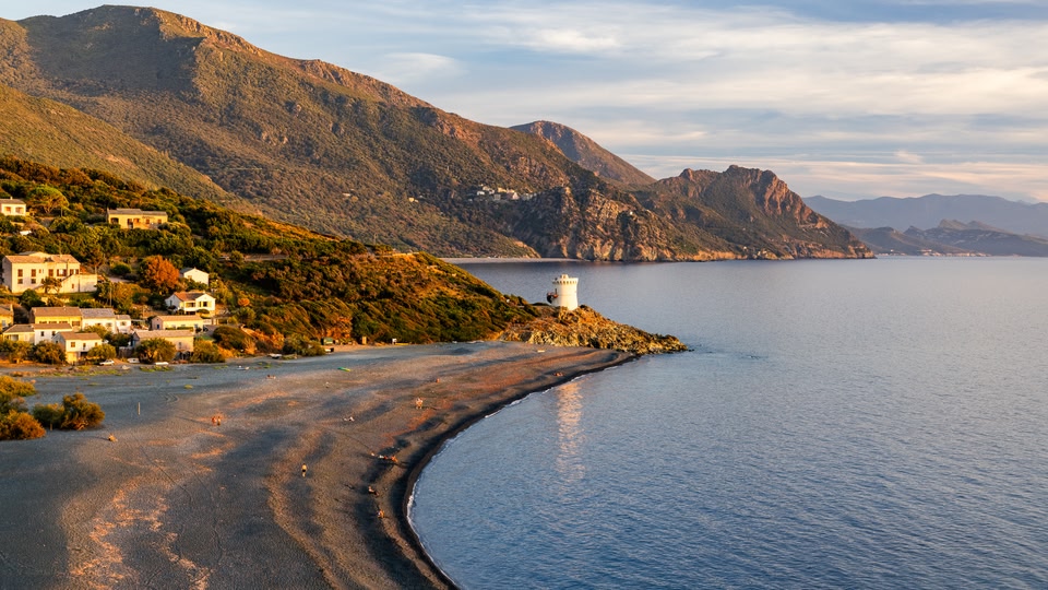 Pebble beach, white round lighthouse on a rocky point and hillside houses at sunset in Cap Corse.