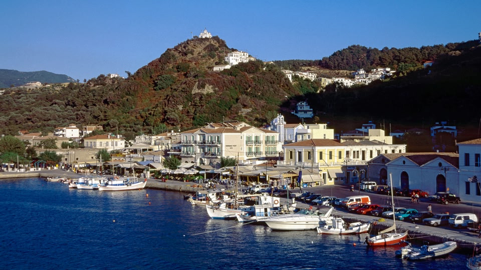 A picturesque coastal town with boats docked along the waterfront, buildings lining the shore, and hills in the background.