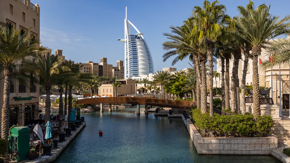A scenic view of the Burj Al Arab in Dubai, showcasing its sail-like design against the clear blue sky, surrounded by modern architecture and palm trees.