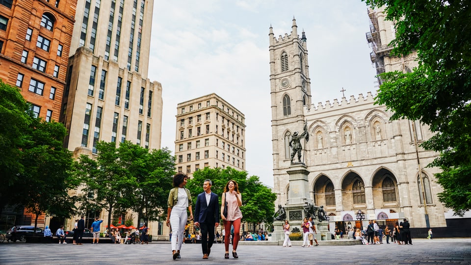 Three people walking through a city square in Montreal with historic buildings and a crowd of people in the background.