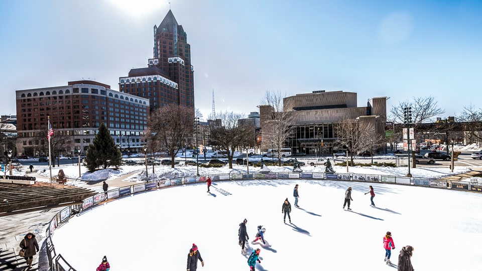 People ice skating on an outdoor rink in an urban area with modern buildings and snow-covered ground.
