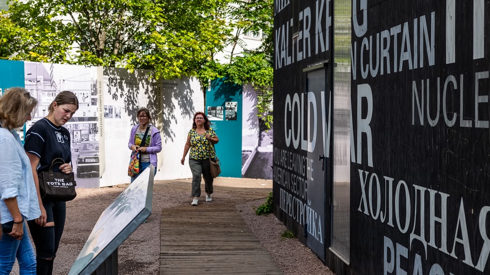 Group of people walking along a path in front of a large, graffiti-covered wall with historical text and images at Checkpoint Charlie in Berlin.