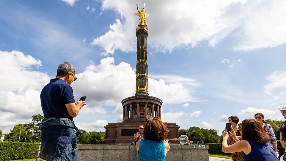 A group of tourists taking photos of the Victory Column in Berlin on a sunny day.