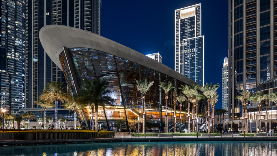 A modern architectural building with a curved design and glass facades, illuminated at night with surrounding palm trees and a reflecting pool.