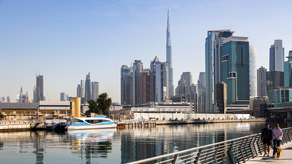 A couple walking their dog along the Dubai Water Canal with a modern city skyline in the background.