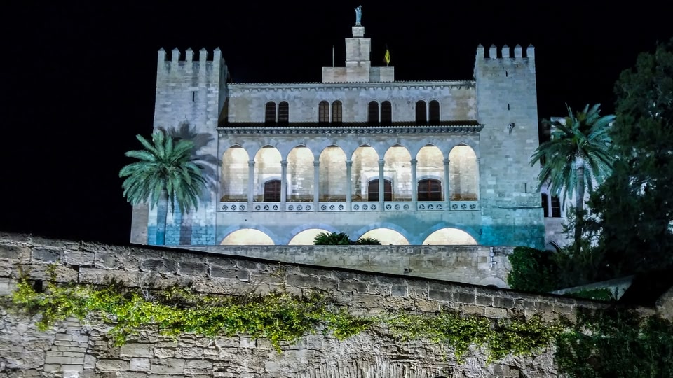 Illuminated historic building at night with stone wall and palm trees