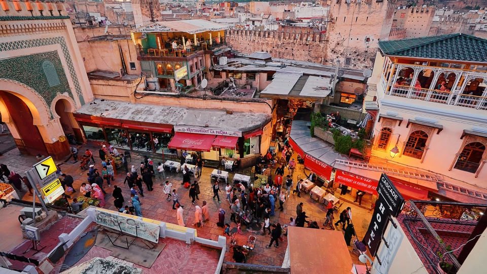 Crowded medina street with market stalls, outdoor tables and tiled rooftops in Fes.