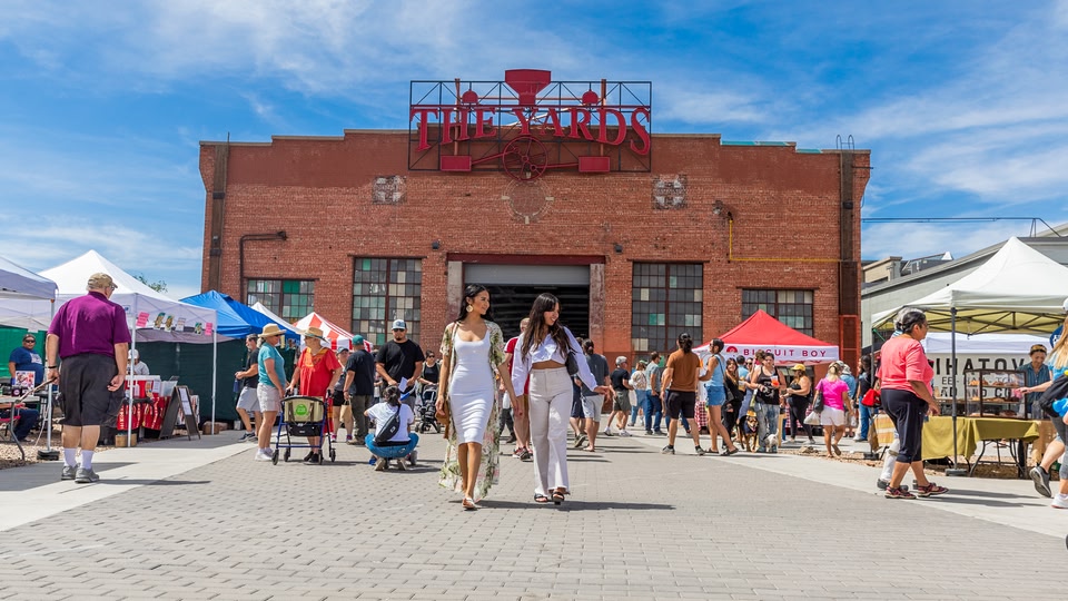 A busy outdoor market with stalls and people walking around under a blue sky.