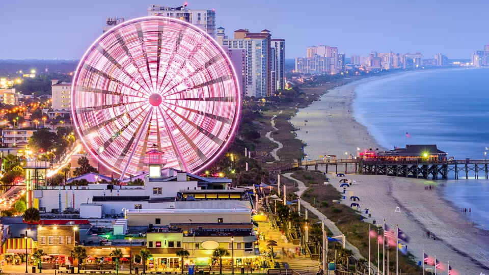 Aerial view of Myrtle Beach at dusk showing a large Ferris wheel, beachfront, and illuminated buildings.