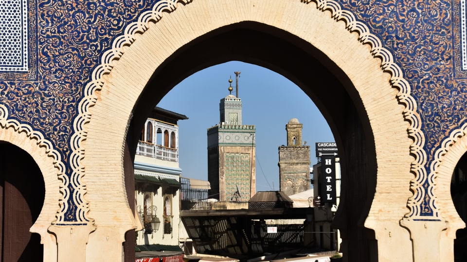 Ornate blue-tiled archway framing a tile-decorated minaret and street buildings in Fes.