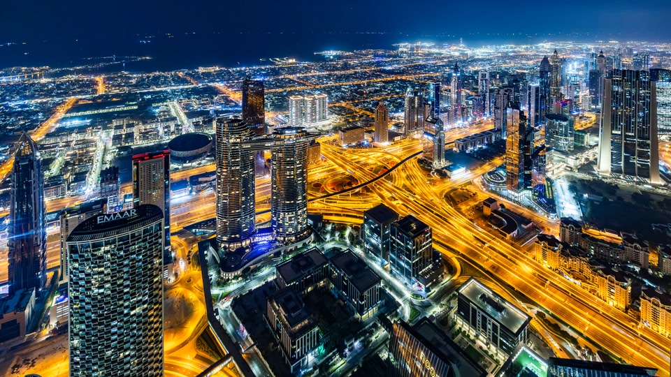 Aerial night view of Dubai's skyline showing multiple illuminated high-rise buildings and busy highways.