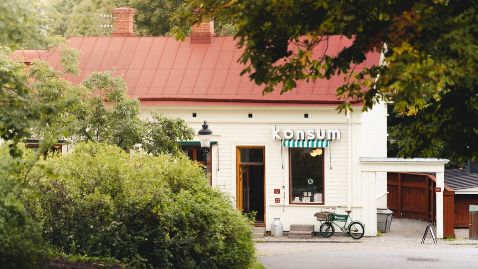 White building with a red roof and a green awning that says 'Konsum'. A bicycle is parked in front of the entrance.
