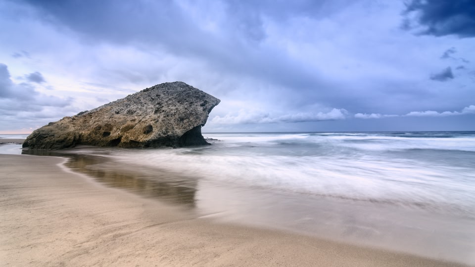 A large rock formation on a sandy beach with crashing waves under a cloudy sky.