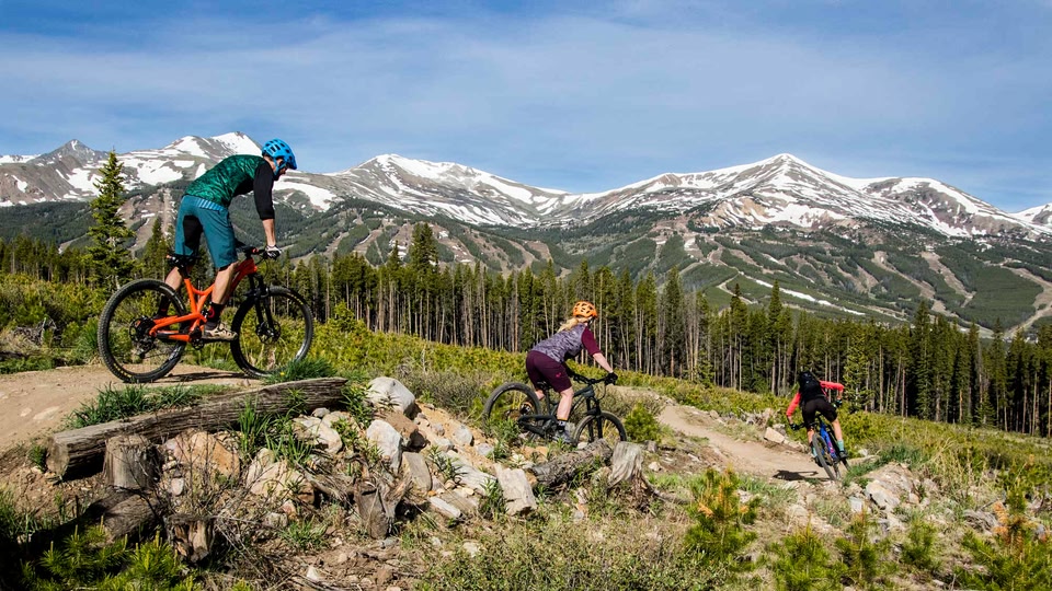 Three cyclists riding mountain bikes on a trail with a scenic mountain range in the background.